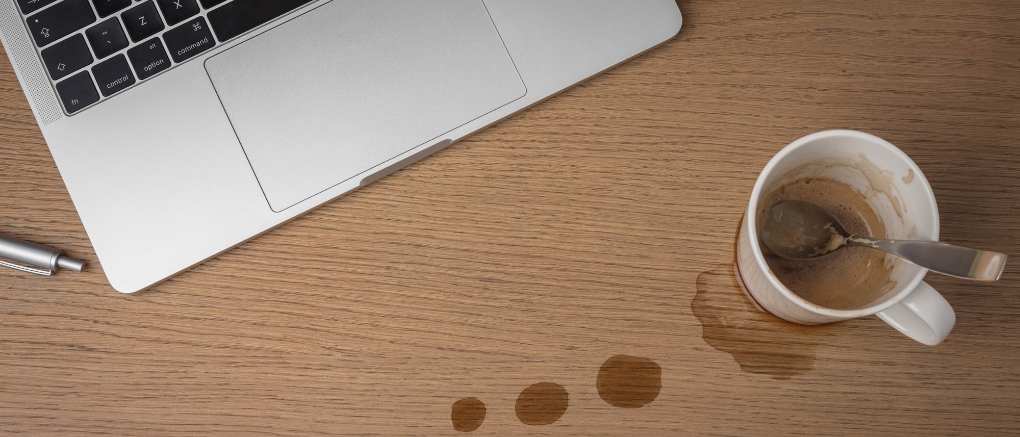 Een laptop en een koffiekopje, er is koffie gemorst op de houten ondergrond
